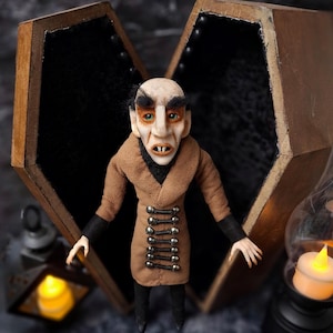 Front view of a handmade Nosferatu art doll standing in front of an open wooden coffin, designed with special fabrics and accessories inside, featuring a cross on top, a unique gothic vampire collectible perfect for spooky art displays, gothic decor, horror themed collections, and dark fantasy setups.