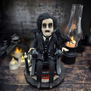 Front view of a handmade Edgar Allan Poe art doll sitting on a rocking chair on a small carpeted stand, with a raven perched nearby, a poseable art doll and unique Poe collectible perfect for literary decor, gothic home accent, and dark fantasy displays.
