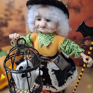 Close-up of the handmade witch art doll from a left angled side view, showing her Halloween-themed outfit, detailed face, the cage with a tiny ghost in one hand, and bat-shaped wand in the other.