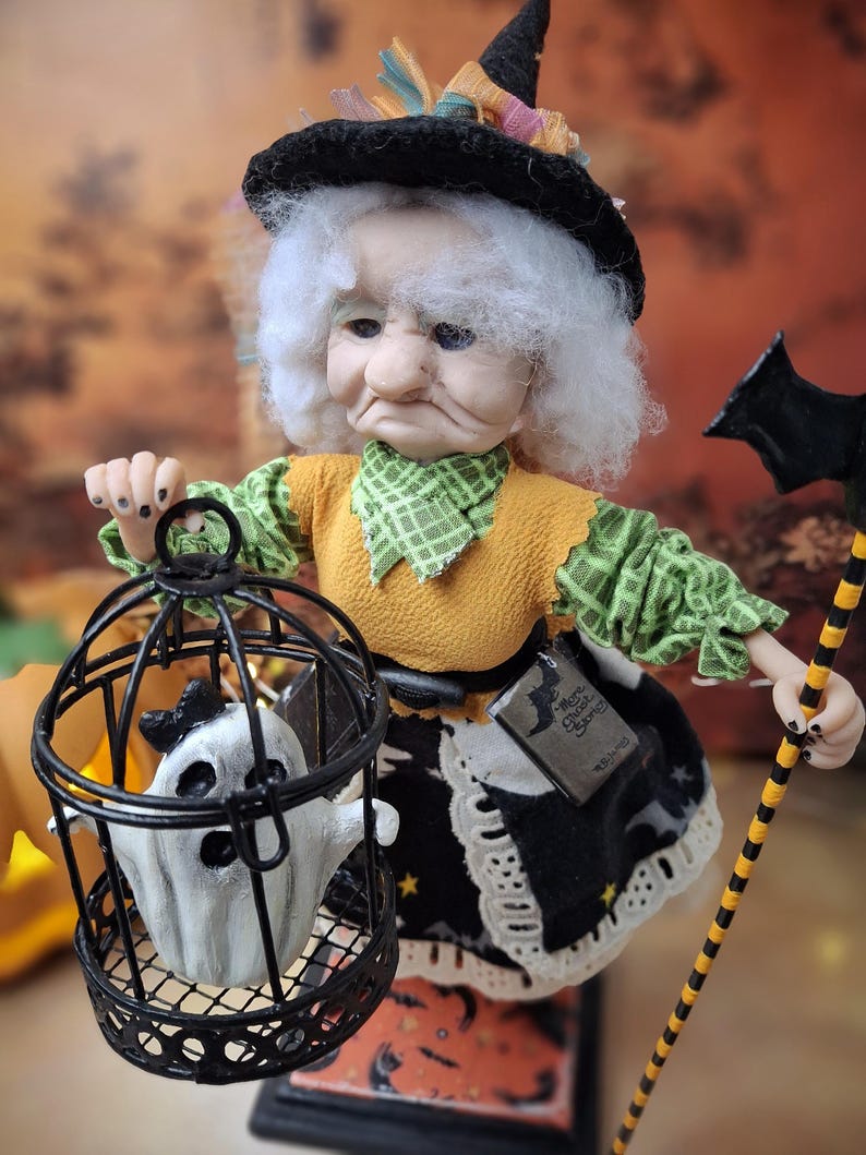 Close-up of the handmade witch art doll from a left angled side view, showing her Halloween-themed outfit, detailed face, the cage with a tiny ghost in one hand, and bat-shaped wand in the other.