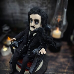 Top-down view from the left of a handmade Edgar Allan Poe art doll sitting on a rocking chair on a small carpeted stand, with a raven perched nearby, a poseable art doll and unique Poe collectible perfect for literary decor, gothic home accent, and dark fantasy displays, highlighting detailed sculpting and painted features.