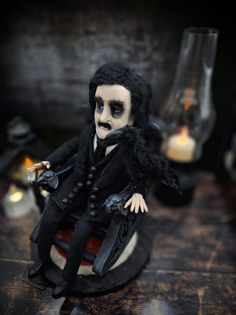 Top-down view from the left of a handmade Edgar Allan Poe art doll sitting on a rocking chair on a small carpeted stand, with a raven perched nearby, a poseable art doll and unique Poe collectible perfect for literary decor, gothic home accent, and dark fantasy displays, highlighting detailed sculpting and painted features.