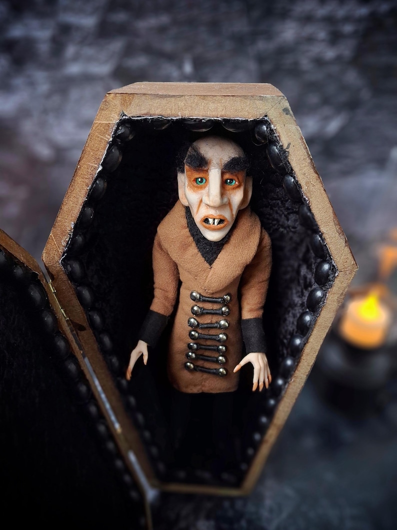 Front view of a handmade Nosferatu art doll sitting inside an open wooden coffin, designed with special fabrics and accessories, featuring a cross on top, a unique gothic vampire collectible perfect for spooky art displays, gothic decor, horror themed collections, and dark fantasy setups, highlighting detailed sculpting and costume features.