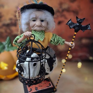 Close-up front view of the handmade witch art doll, slightly leaning to the right, wearing a Halloween-themed outfit and holding a cage with a tiny ghost in one hand and a bat-shaped wand in the other.
