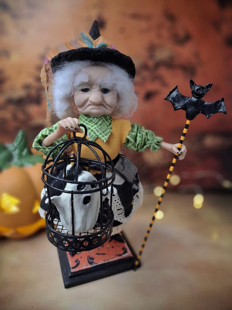 Close-up front view of the handmade witch art doll, slightly leaning to the right, wearing a Halloween-themed outfit and holding a cage with a tiny ghost in one hand and a bat-shaped wand in the other.