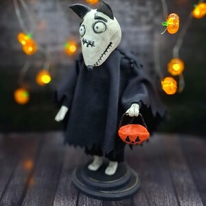 Handmade Sparky Art Doll wearing a black hoodie Halloween-style outfit, holding a pumpkin-shaped trick-or-treat box. Viewed from a low left diagonal angle. Whimsical and spooky Halloween home decor, unique collectible dog doll