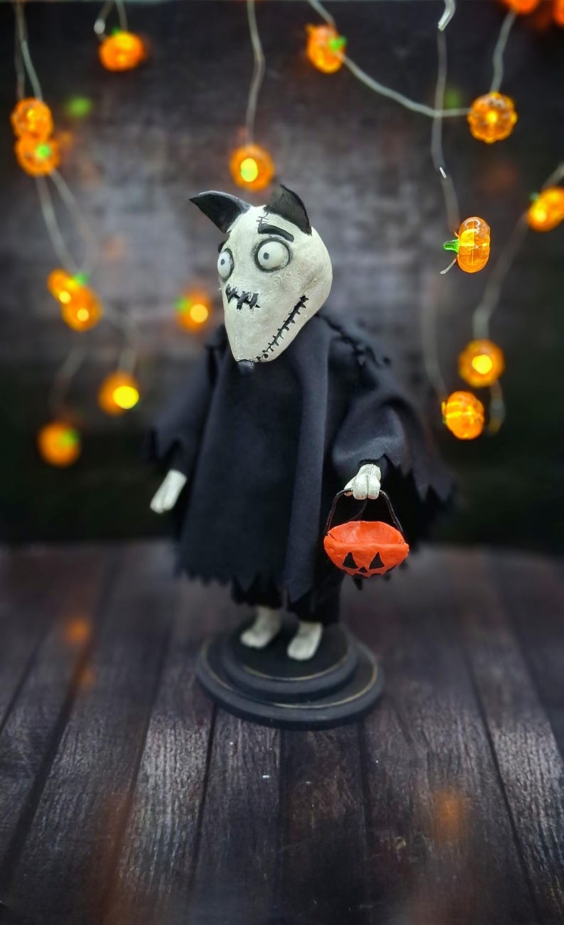 Handmade Sparky Art Doll wearing a black hoodie Halloween-style outfit, holding a pumpkin-shaped trick-or-treat box. Viewed from a low left diagonal angle. Whimsical and spooky Halloween home decor, unique collectible dog doll
