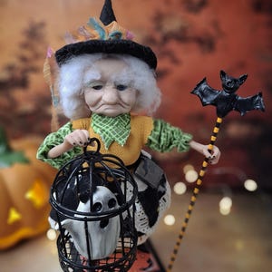 Top-down view of the handmade witch art doll angled slightly to the right, wearing a Halloween-themed outfit and holding a cage with a tiny ghost in one hand and a bat-shaped wand in the other.