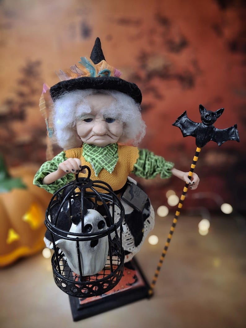 Top-down view of the handmade witch art doll angled slightly to the right, wearing a Halloween-themed outfit and holding a cage with a tiny ghost in one hand and a bat-shaped wand in the other.