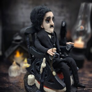 Profile view from the right of a handmade Edgar Allan Poe art doll sitting on a rocking chair on a small carpeted stand, with a raven perched nearby, a poseable art doll and unique Poe collectible perfect for literary decor, gothic home accent, and dark fantasy displays, showing detailed sculpting and painted features.