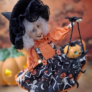 Right-angled view of the handmade witch art doll, turned slightly more to the right, holding a tiny pumpkin skeleton puppet and wearing a cute Halloween-themed outfit with spooky whimsical details.