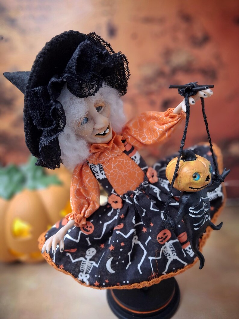 Right-angled view of the handmade witch art doll, turned slightly more to the right, holding a tiny pumpkin skeleton puppet and wearing a cute Halloween-themed outfit with spooky whimsical details.