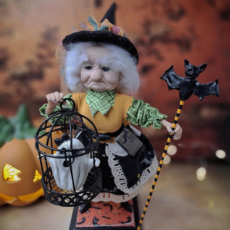 Front view of a handmade witch art doll wearing a Halloween-themed outfit, holding a cage with a tiny ghost in one hand and a bat-shaped wand in the other, looking at the cage.