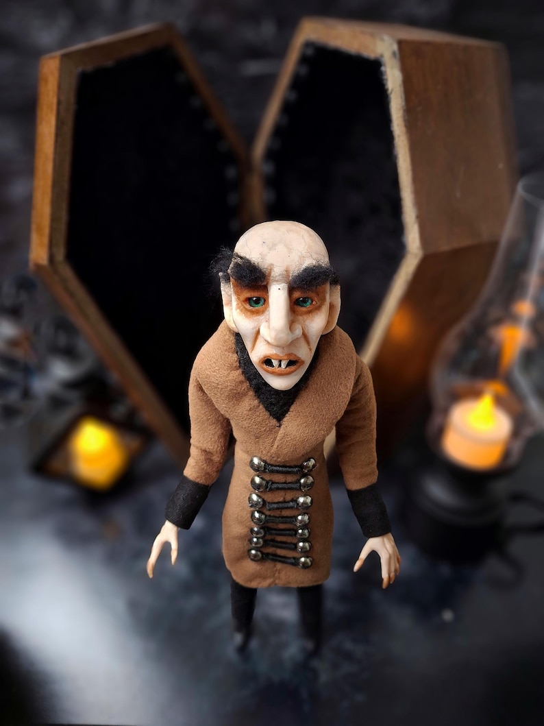 Top-down front close-up view of a handmade Nosferatu art doll standing in front of an open wooden coffin, designed with special fabrics and accessories, featuring a cross on top, a unique gothic vampire collectible perfect for spooky art displays, gothic decor, horror themed collections, and dark fantasy setups, highlighting detailed sculpting and costume features.