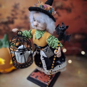 Top-down view of the handmade witch art doll from the left side profile, showing her Halloween-themed outfit, the cage with a tiny ghost in one hand, and the bat-shaped wand in the other.
