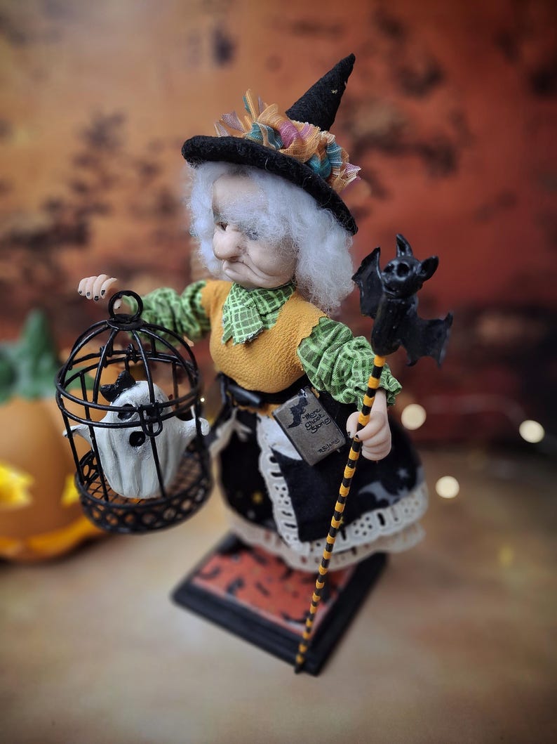 Top-down view of the handmade witch art doll from the left side profile, showing her Halloween-themed outfit, the cage with a tiny ghost in one hand, and the bat-shaped wand in the other.