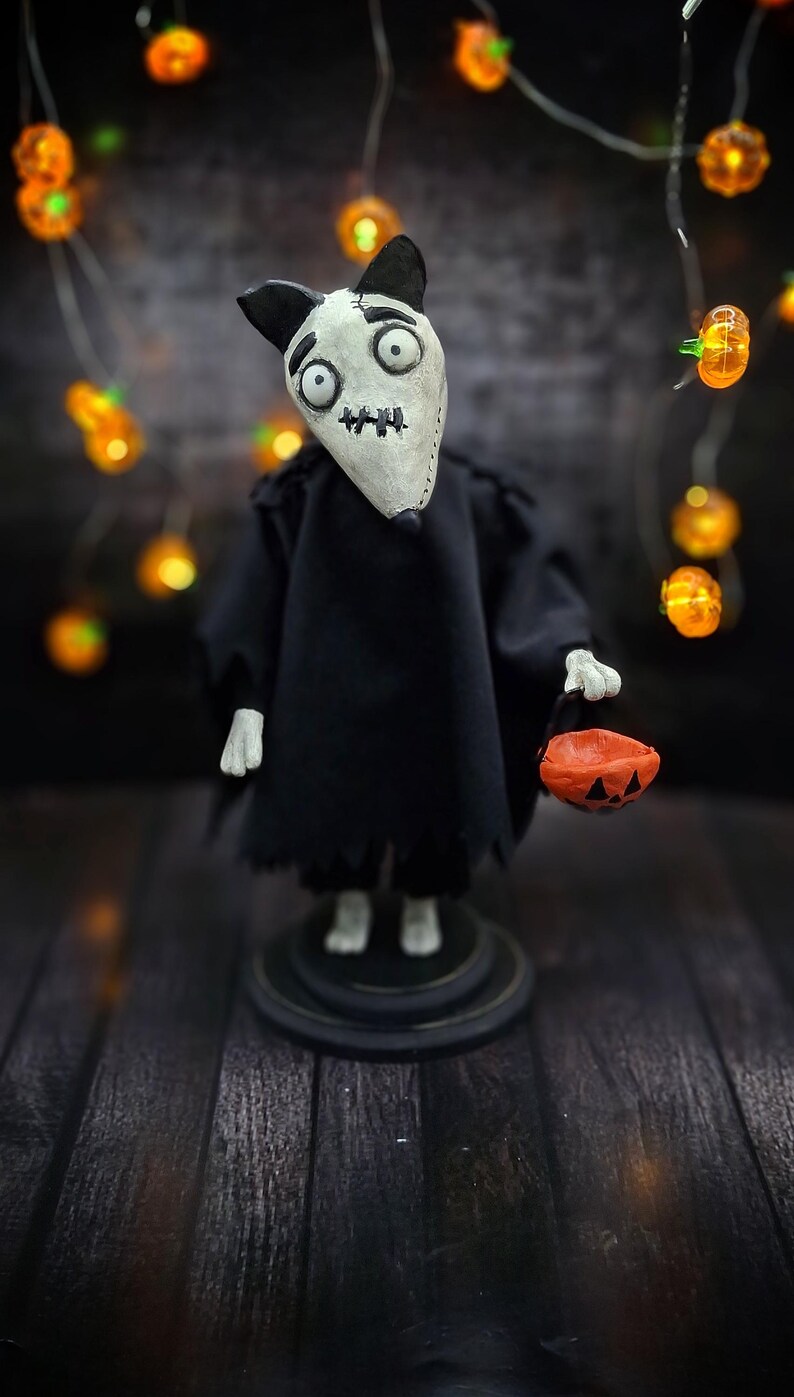 Handmade Sparky Art Doll wearing a black hoodie Halloween-style outfit, holding a pumpkin-shaped trick-or-treat box. Standing straight, looking surprised directly at the viewer. Whimsical and spooky Halloween home decor, unique collectible dog doll