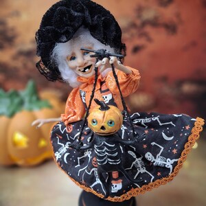 Slightly left-leaning view of the handmade witch art doll holding a tiny pumpkin skeleton puppet, showing her adorable Halloween outfit and detailed spooky cute design.