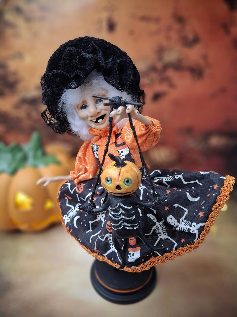 Slightly left-leaning view of the handmade witch art doll holding a tiny pumpkin skeleton puppet, showing her adorable Halloween outfit and detailed spooky cute design.