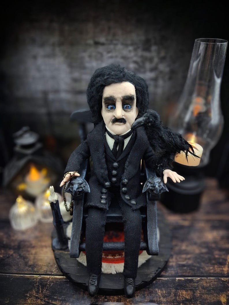Close-up front view of a handmade Edgar Allan Poe art doll sitting on a rocking chair on a small carpeted stand, with a raven perched nearby, a poseable art doll and unique Poe collectible perfect for literary decor, gothic home accent, and dark fantasy displays, highlighting detailed sculpting and painted features.