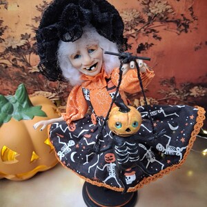 Medium close-up view of the handmade witch art doll, slightly leaning to the left, holding a tiny pumpkin skeleton puppet and wearing a cute Halloween-themed outfit with whimsical spooky details.