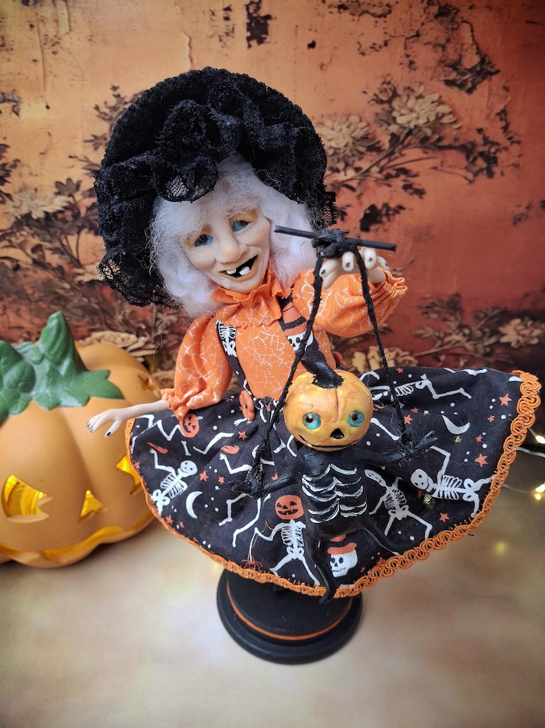 Medium close-up view of the handmade witch art doll, slightly leaning to the left, holding a tiny pumpkin skeleton puppet and wearing a cute Halloween-themed outfit with whimsical spooky details.