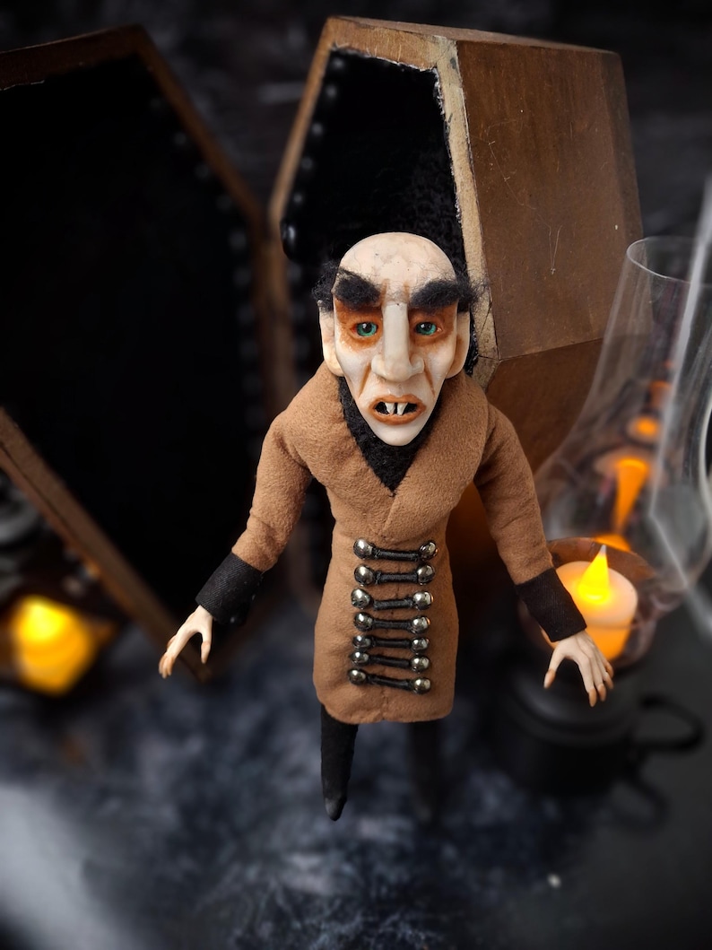 Front-right view of a handmade Nosferatu art doll standing to the right in front of an open wooden coffin, designed with special fabrics and accessories, featuring a cross on top, a unique gothic vampire collectible perfect for spooky art displays, gothic decor, horror themed collections, and dark fantasy setups, highlighting detailed sculpting and costume features.