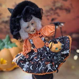 Top-down close-up view of the handmade witch art doll, showing her detailed face, Halloween-themed outfit, and the tiny pumpkin skeleton puppet she holds.
