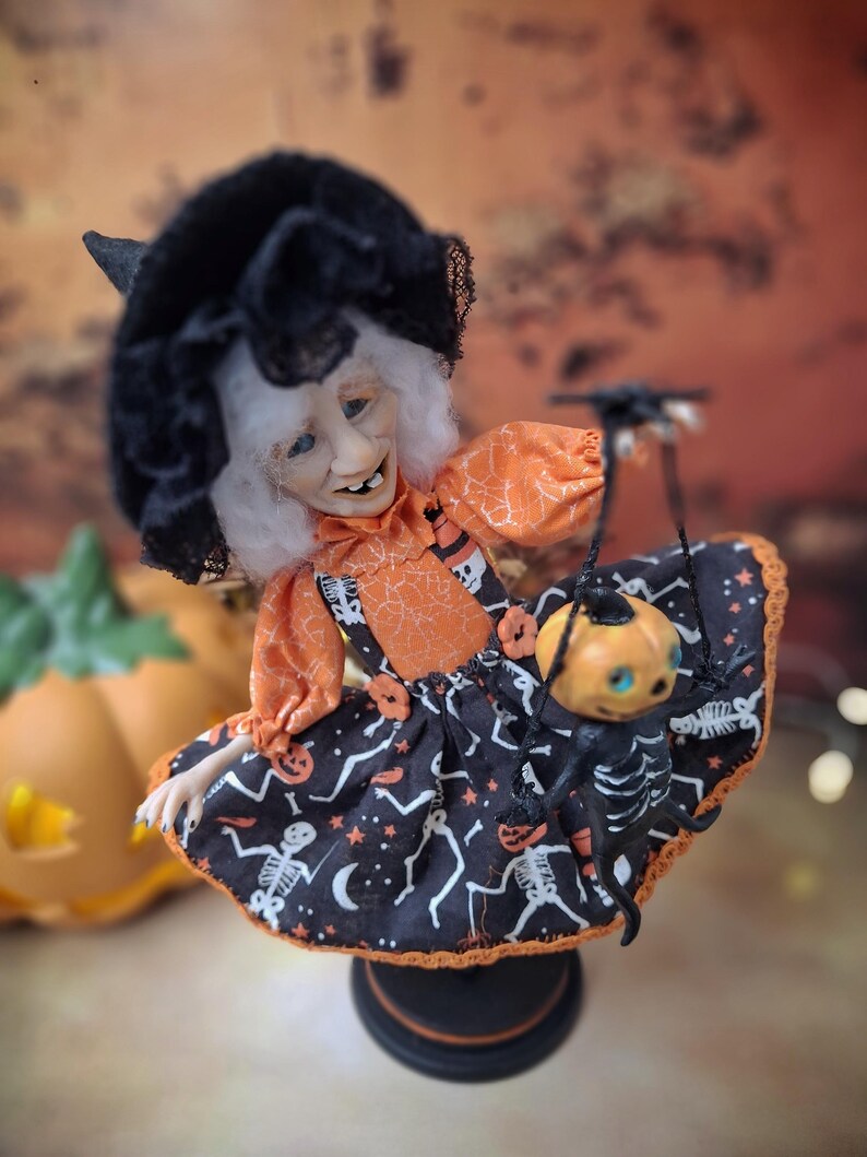 Top-down close-up view of the handmade witch art doll, showing her detailed face, Halloween-themed outfit, and the tiny pumpkin skeleton puppet she holds.