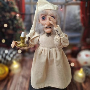 A close-up front view of the Ebenezer Scrooge Art Doll, showing one hand resting near his face. The detailed hand-painted expression, textured vintage-inspired outfit, and subtle holiday accents are clearly visible, emphasizing the doll’s storybook charm and whimsical character.