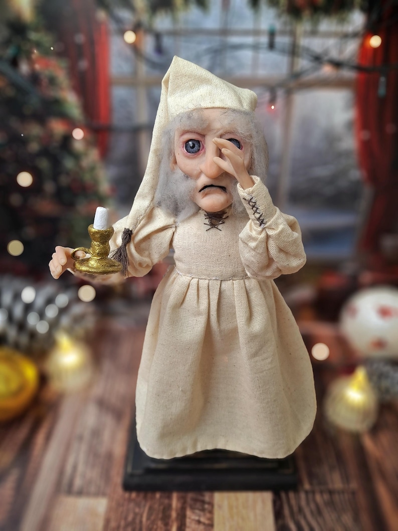A close-up front view of the Ebenezer Scrooge Art Doll, showing one hand resting near his face. The detailed hand-painted expression, textured vintage-inspired outfit, and subtle holiday accents are clearly visible, emphasizing the doll’s storybook charm and whimsical character.