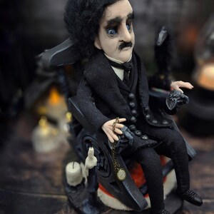 Top-down view from the right of a handmade Edgar Allan Poe art doll sitting on a rocking chair on a small carpeted stand, with a raven nearby, a poseable art doll and unique Poe collectible perfect for literary decor, gothic home accent, and dark fantasy displays, highlighting detailed sculpting and painted features.