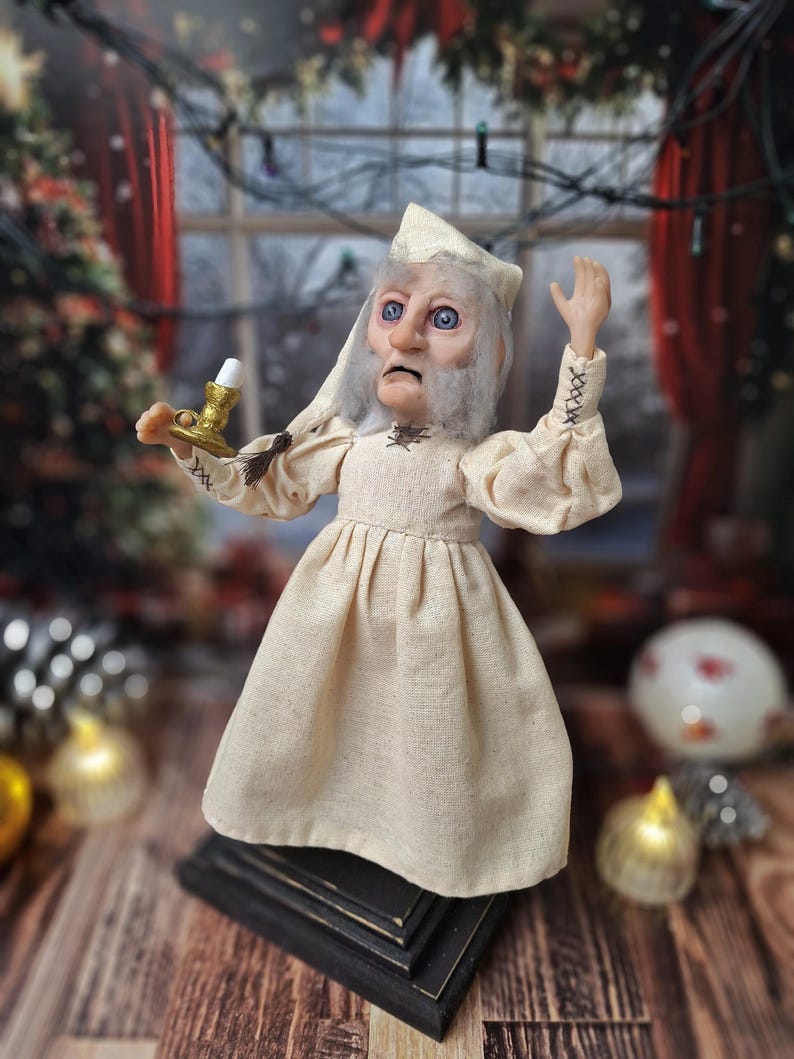 A left-angled view of the Ebenezer Scrooge Art Doll looking slightly upward, showcasing the intricate details of his hand-painted face and vintage-inspired outfit. The mixed-media textures and subtle holiday accents highlight the whimsical, storybook charm of the character.