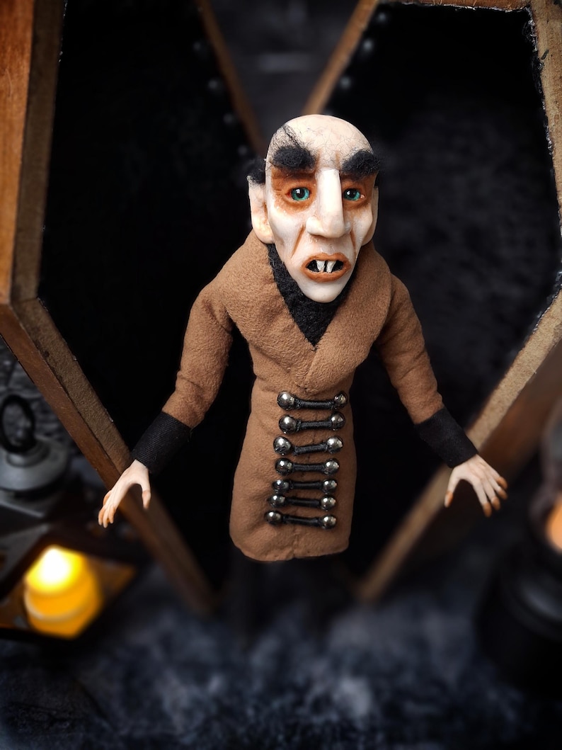 Front view of a handmade Nosferatu art doll standing in front of an open wooden coffin, slightly tilted to the right, designed with special fabrics and accessories, featuring a cross on top, a unique gothic vampire collectible perfect for spooky art displays, gothic decor, horror themed collections, and dark fantasy setups, highlighting detailed sculpting and costume features.