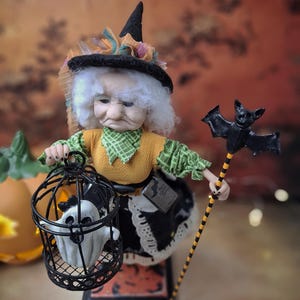 Top-down view of a handmade witch art doll in a Halloween-themed outfit, holding a cage with a tiny ghost in one hand and a bat-shaped wand in the other, looking at the cage.