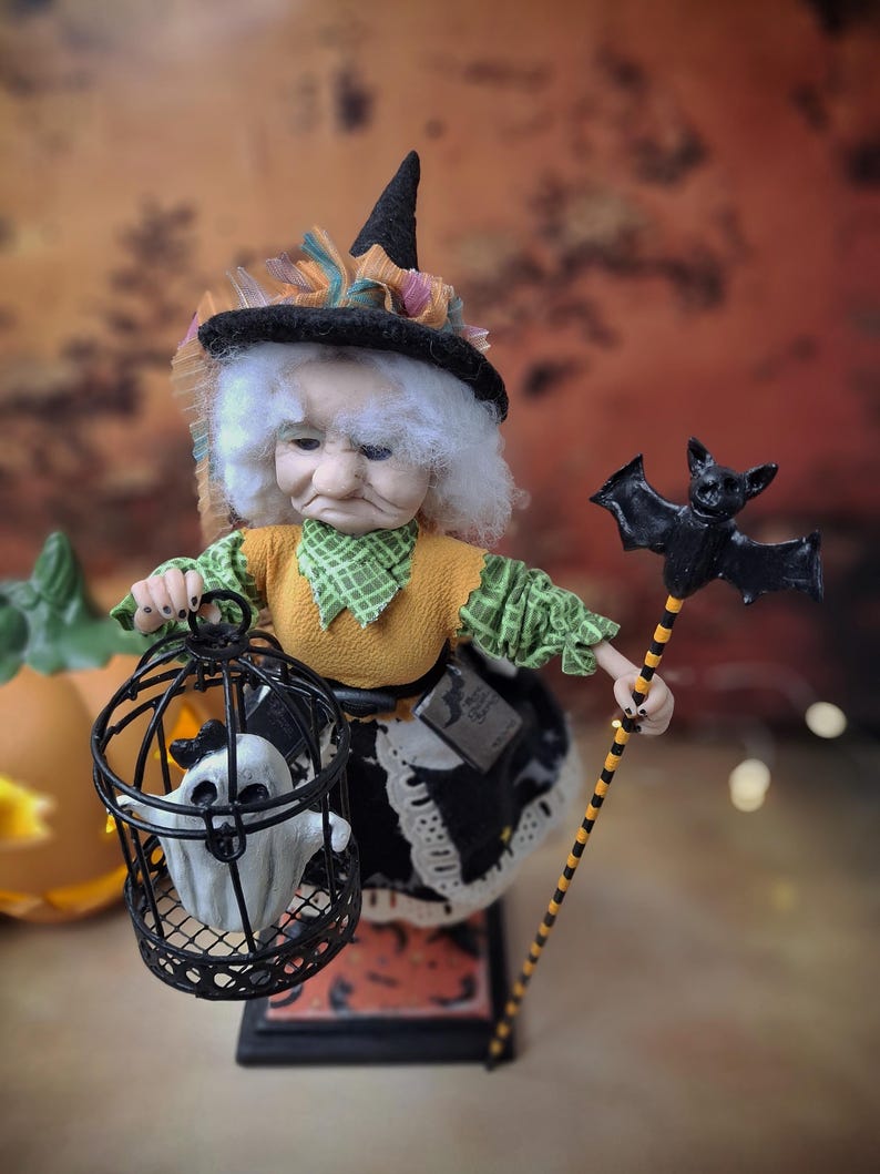 Top-down view of a handmade witch art doll in a Halloween-themed outfit, holding a cage with a tiny ghost in one hand and a bat-shaped wand in the other, looking at the cage.