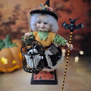 Top-down view of the handmade witch art doll angled slightly to the left, wearing a Halloween-themed outfit and holding a cage with a tiny ghost in one hand and a bat-shaped wand in the other.