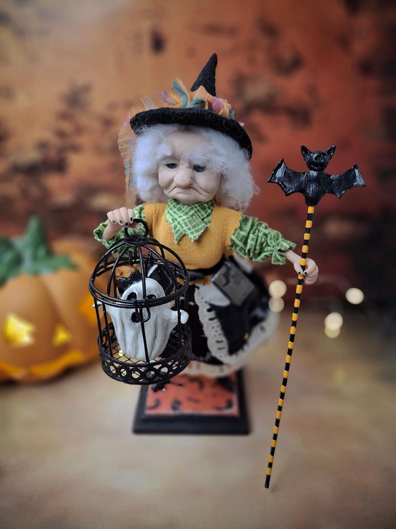 Top-down view of the handmade witch art doll angled slightly to the left, wearing a Halloween-themed outfit and holding a cage with a tiny ghost in one hand and a bat-shaped wand in the other.