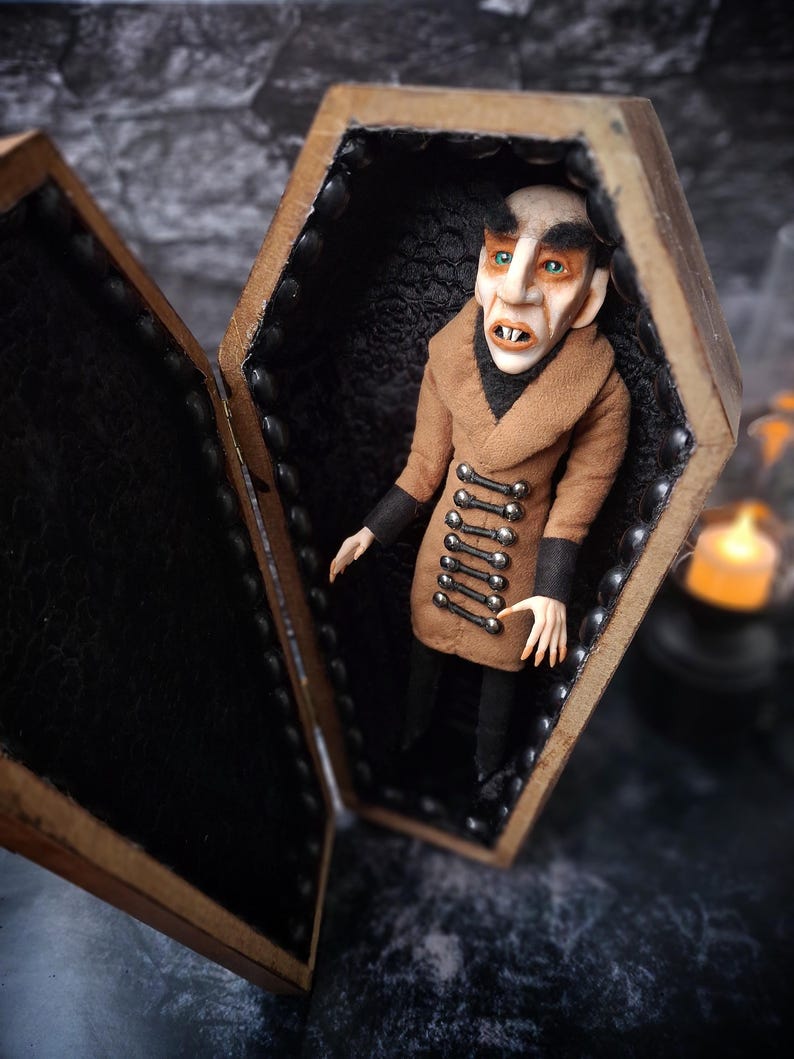 Front-left view of a handmade Nosferatu art doll sitting inside an open wooden coffin, facing left, designed with special fabrics and accessories, featuring a cross on top, a unique gothic vampire collectible perfect for spooky art displays, gothic decor, horror themed collections, and dark fantasy setups, highlighting detailed sculpting and costume features.