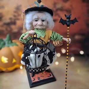 Low-angle view from the right of the handmade witch art doll, wearing a Halloween-themed outfit and holding a cage with a tiny ghost in one hand and a bat-shaped wand in the other.
