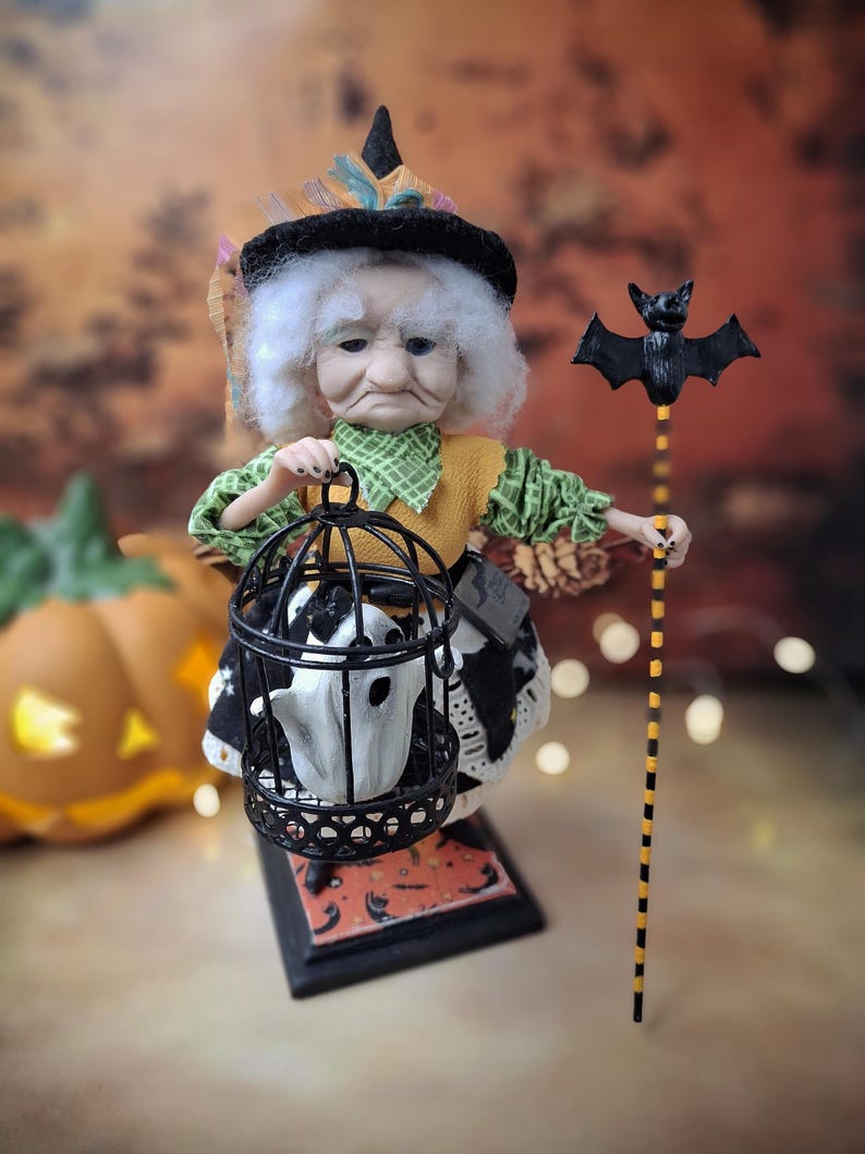 Low-angle view from the right of the handmade witch art doll, wearing a Halloween-themed outfit and holding a cage with a tiny ghost in one hand and a bat-shaped wand in the other.