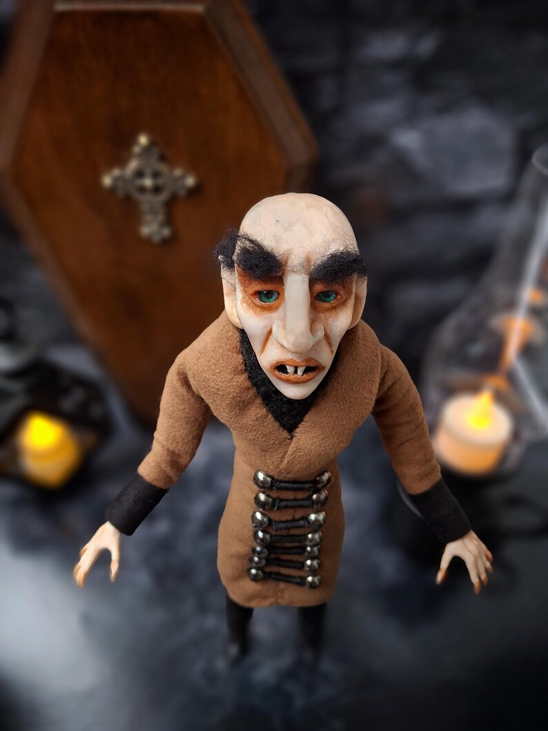 Top-down front close-up view of a handmade Nosferatu art doll standing in front of a closed wooden coffin, designed with special fabrics and accessories, featuring a cross on top, a unique gothic vampire collectible perfect for spooky art displays, gothic decor, horror themed collections, and dark fantasy setups, highlighting detailed sculpting and costume features.