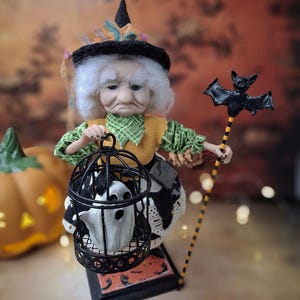 Distant top-down view of the handmade witch art doll angled slightly to the right, showing her full Halloween-themed outfit, the cage with a tiny ghost in one hand, and the bat-shaped wand in the other.