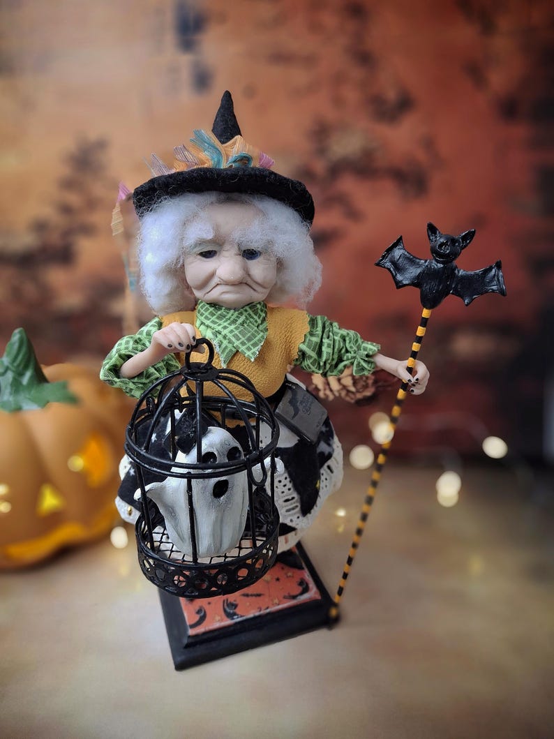 Distant top-down view of the handmade witch art doll angled slightly to the right, showing her full Halloween-themed outfit, the cage with a tiny ghost in one hand, and the bat-shaped wand in the other.