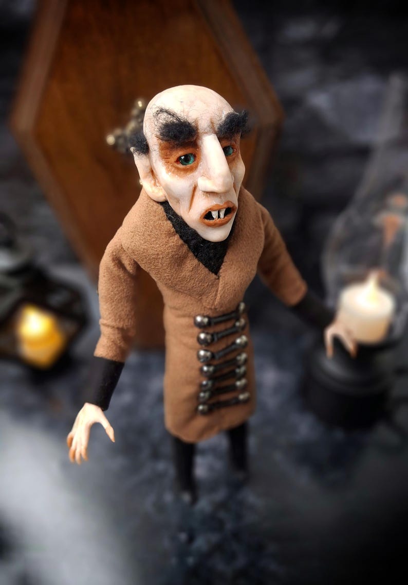 Top-right diagonal view of a handmade Nosferatu art doll standing in front of a closed wooden coffin, designed with special fabrics and accessories, featuring a cross on top, a unique gothic vampire collectible perfect for spooky art displays, gothic decor, horror themed collections, and dark fantasy setups, highlighting detailed sculpting and costume features.