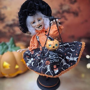 Front view of the handmade witch art doll standing upright, featuring her whimsical Halloween outfit, detailed facial features, and holding a tiny handmade pumpkin skeleton puppet