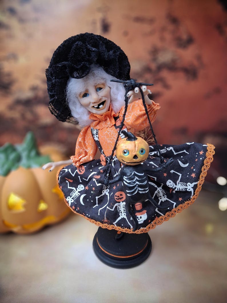 Front view of the handmade witch art doll standing upright, featuring her whimsical Halloween outfit, detailed facial features, and holding a tiny handmade pumpkin skeleton puppet