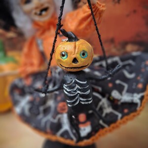 Close-up of the tiny handmade pumpkin skeleton puppet held by the witch art doll, showing detailed sculpting and cute spooky Halloween design elements.