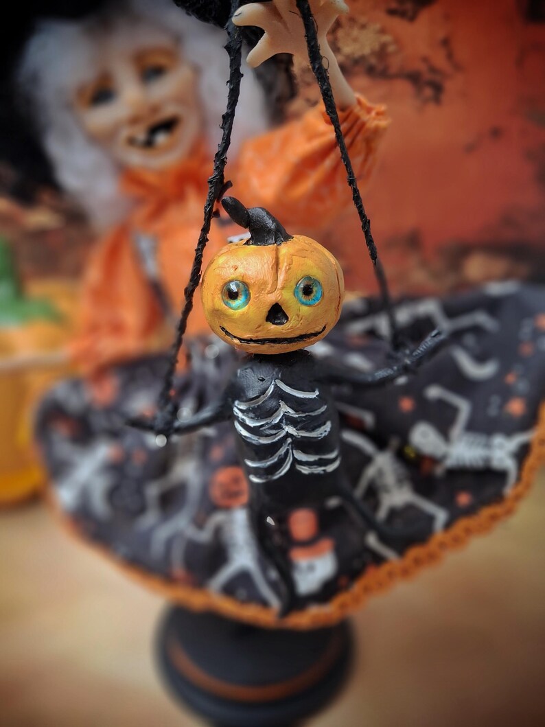 Close-up of the tiny handmade pumpkin skeleton puppet held by the witch art doll, showing detailed sculpting and cute spooky Halloween design elements.