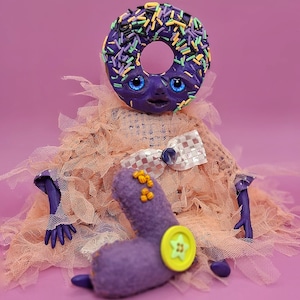 Puede incluir: Una criatura caprichosa en forma de donut morado con ojos azules y chispas de colores. Tiene una pajarita, un botón y un cuerpo suave y texturizado. El fondo es de color rosa liso.
